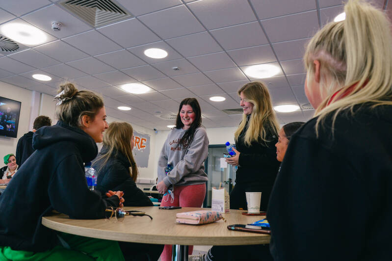 A group of SRC students are smiling and chatting round a table in a relaxed casual seating area. There are coffee cups and mobile phones on the table and in their hands.