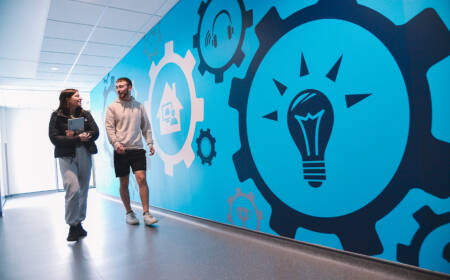 Two students walking along corridor, the wall is bright blue with large decals of lightbulbs, cogs and other tech related symbols