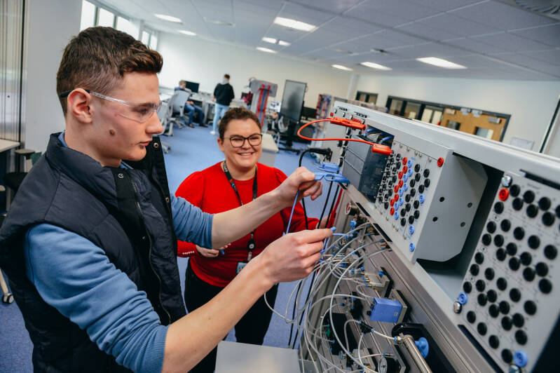 An SRC student is working with a large electronic system in an electronic engineering lab while a member of SRC staff looks on, smiling. They are both wearing safety glasses.